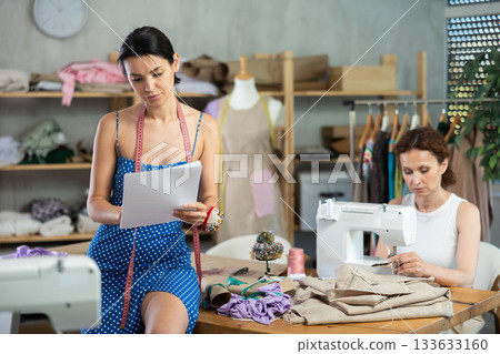 Woman designer draw dress sketch on paper, female tailor sewing in background Woman designer draw dress sketch on paper, female tailor sewing in background 133633160