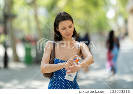 Young brunette girl applying sunscreen to her skin while walking in city 133633216