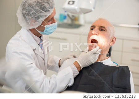 Man doctor dentist in white coat examines oral cavity of mature male patient in dentistry. 133633218