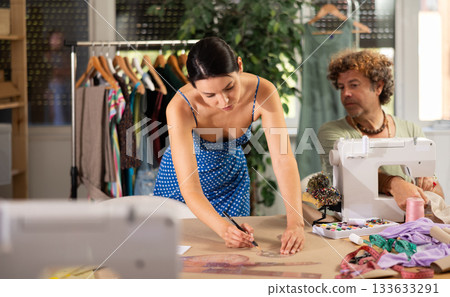 Woman tailor draw dress pattern of product on paper using curve ruler, man sew in background Woman tailor draw dress pattern of product on paper using curve ruler, man sew in background 133633291