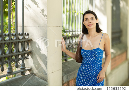 Cheerful brunette girl in blue dress walking along the street among architecture with bag 133633292