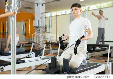 Guy doing exercises with straps in arms on Pilates reformer Guy doing exercises with straps in arms on Pilates reformer 133633317