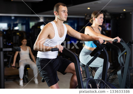 Sportive young couple doing air bike exercises in well-equipped gym Sportive young couple doing air bike exercises in well-equipped gym 133633386