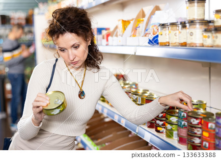 In Russian goods store, woman choose canned sprats in oil 133633388