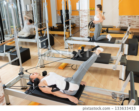 Man works out in gym and performs set of exercises to stretch leg muscles with help of reformer 133633433