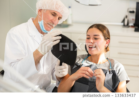 Senior man specialist holds mirror in front of girl patient and suggests evaluating effect of treatment 133633437
