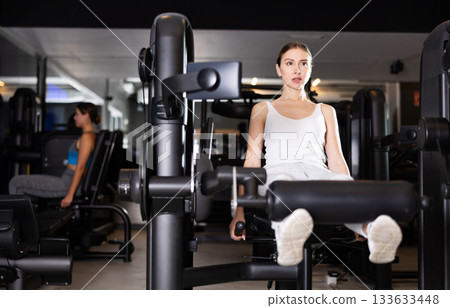 Slender athletic girl work out muscles of legs and thighs on simulator in fitness club 133633448