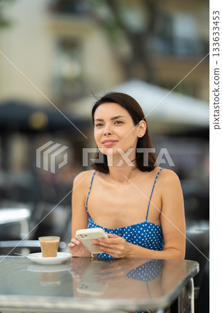 Brunette texting on smartphone screen during breakfast on cafe terrace. Drinking coffee 133633453