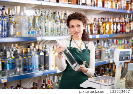 Woman salesperson in alcohol department of Russian store stand with vodka near showcase. Woman salesperson in alcohol department of Russian store stand with vodka near showcase. 133633467