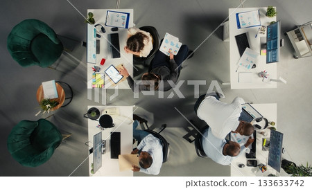 Top down view of corporate employees study business reports and analytics. Colleagues share insights on forecasting, documentation, and targets to improve growth strategy. Top down view of corporate employees study business reports and analytics. Colleagues share insights on forecasting, documentation, and targets to improve growth strategy. 133633742
