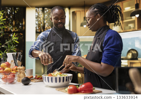 African american man seasoning the salad with mix of spices from a jar, pouring condiments for a tasty vegetarian meal. Smiling woman mixing fresh vegetables together, healthy lifestyle. African american man seasoning the salad with mix of spices from a jar, pouring condiments for a tasty vegetarian meal. Smiling woman mixing fresh vegetables together, healthy lifestyle. 133633914