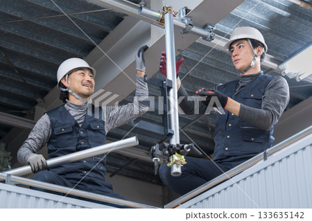 施工場景包括將單一管道固定在靠近天花板的高架鷹架上。圖片中可以看到建築工人正在用扳手擰緊管道。 施工場景包括將單一管道固定在靠近天花板的高架鷹架上。圖片中可以看到建築工人正在用扳手擰緊管道。 133635142