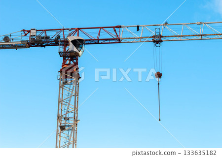 Close-up Red-and-white Tower Crane on Construction Site against Blue sky on Sunny day. Column Crane Boom arrangement with Hook suspension and Counterweight at Ends. Close-up Red-and-white Tower Crane on Construction Site against Blue sky on Sunny day. Column Crane Boom arrangement with Hook suspension and Counterweight at Ends. 133635182