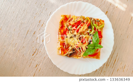 Slice of pizza with cheese, vegetables and basil on a white plate standing on a wooden table. 133636349