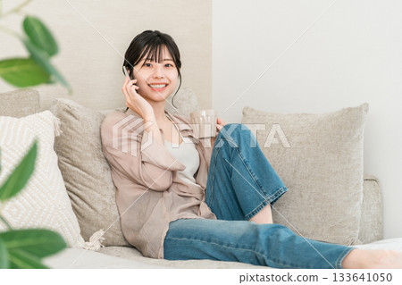 日本女子一邊喝熱飲一邊用手機打電話(家中,客廳,面帶微笑) 日本女子一邊喝熱飲一邊用手機打電話(家中,客廳,面帶微笑) 133641050