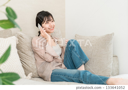 日本女子一邊喝熱飲一邊用手機打電話（家中，客廳，面帶微笑） 133641052