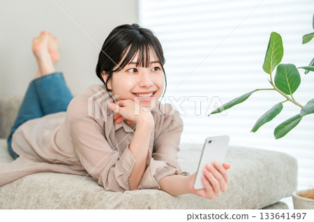 A young Asian woman lying on the sofa and relaxing while looking at her smartphone 133641497