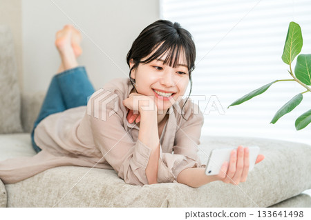 一位年輕的亞洲女性躺在沙發上放鬆地看著她的智慧型手機 133641498