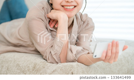 一位年輕的亞洲女性躺在沙發上放鬆地看著她的智慧型手機 133641499