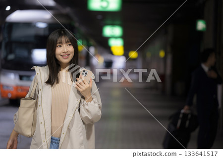一位女性在夜間的城市車站或公車樞紐站看著智慧型手機，這是她通勤回家途中展現的都市生活場景。 133641517