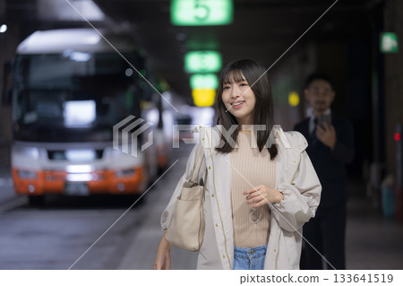 一位女性在夜間的城市車站或公車樞紐站看著智慧型手機，這是她通勤回家途中展現的都市生活場景。 133641519