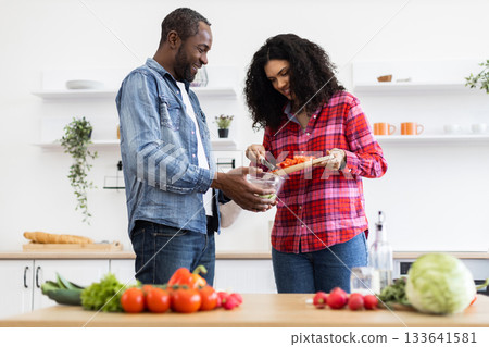 A smiling couple prepares a meal together in a bright, modern kitchen, chopping vegetables. 133641581