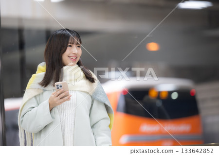 一位女士戴著可愛的圍巾，手持智慧型手機，出現在火車站台或汽車站，這是她冬季新年假期回程的場景。 133642852