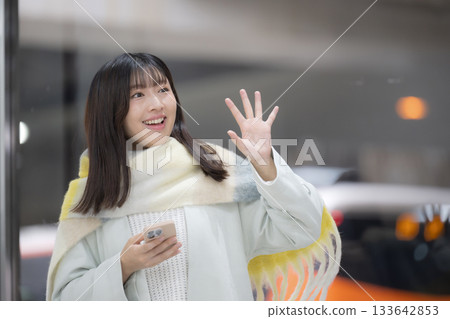 A woman wearing a cute scarf and holding a smartphone at a train station platform or bus terminal in winter, waving goodbye 133642853