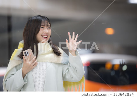 A woman wearing a cute scarf and holding a smartphone at a train station platform or bus terminal in winter, waving goodbye 133642854