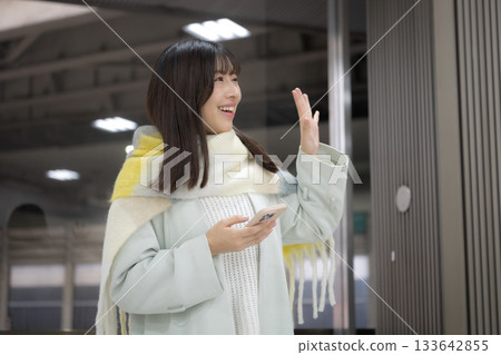 A woman wearing a cute scarf and holding a smartphone at a train station platform or bus terminal in winter, waving goodbye 133642855