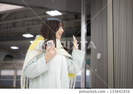 A woman wearing a cute scarf and holding a smartphone at a train station platform or bus terminal in winter, waving goodbye 133642856