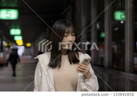 A woman's commute home scene and urban lifestyle, looking at her smartphone at a city station, airport, or bus terminal at night A woman's commute home scene and urban lifestyle, looking at her smartphone at a city station, airport, or bus terminal at night 133642858