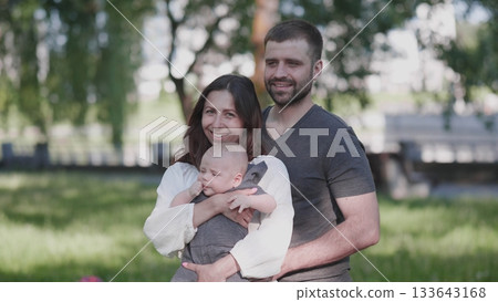 A joyful family stands together in a park, with the mother holding their baby and the father smiling beside them, all basking in the warm sunshine. 133643168