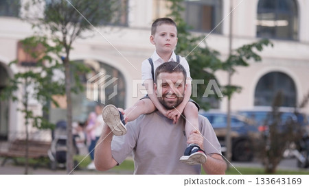 A father joyfully carries his son on his shoulders in a lively park setting filled with trees and families enjoying a sunny afternoon together. 133643169