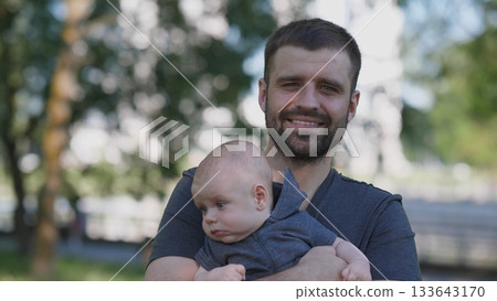 A father enjoys quality time with his baby in a park, surrounded by trees and natural light, reflecting a moment of joy and connection on a sunny day. 133643170