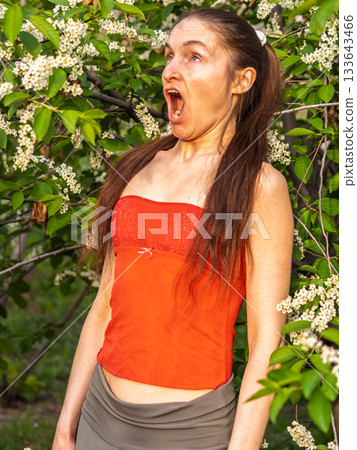 Young woman sneezes in the park against the background of a flowering tree. Allergy to pollen concept. Young woman sneezes in the park against the background of a flowering tree. Allergy to pollen concept. 133643466