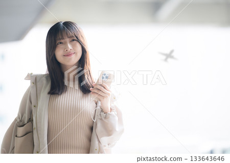 Photo of a woman holding a smartphone at an airport or public transport terminal with an airplane in the background Photo of a woman holding a smartphone at an airport or public transport terminal with an airplane in the background 133643646