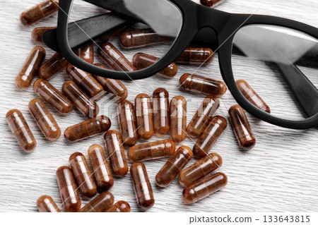Black rimmed eyeglasses on neutral grey plank board with brown supplement capsules sprinkled across the aged lumber texture Black rimmed eyeglasses on neutral grey plank board with brown supplement capsules sprinkled across the aged lumber texture 133643815