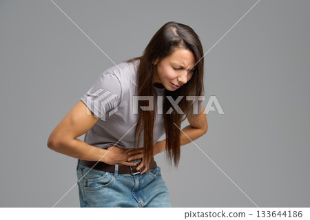 Woman bending forward and holding abdomen in pain, studio scene on neutral grey Woman bending forward and holding abdomen in pain, studio scene on neutral grey 133644186