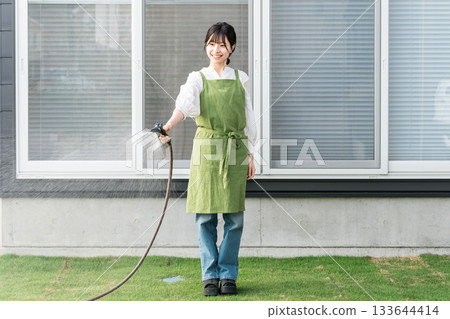 Asian woman wearing an apron watering the lawn in her garden 133644414