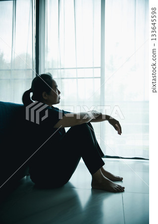 A lonely woman sits on the floor by window looking outside in dark room. Concept of depression, anxiety, sadness, loneliness, mental health problem, emotional struggle and hope for better life. A lonely woman sits on the floor by window looking outside in dark room. Concept of depression, anxiety, sadness, loneliness, mental health problem, emotional struggle and hope for better life. 133645598