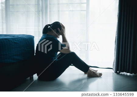 A lonely woman sits on the floor by window with hand on head in dark room. Concept of depression, sadness, anxiety, stress, mental health problem, loneliness and emotional struggle in life. 133645599