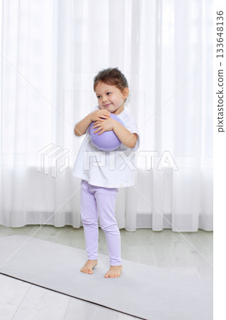 Toddler girl embracing purple pilates ball, standing on a yoga mat in a bright room 133648136