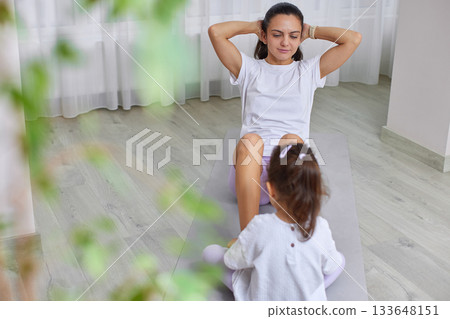Mother doing crunches on a yoga mat with her daughter watching nearby 133648151