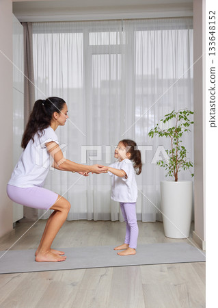 Mother and daughter exercising together on a yoga mat, doing a squat 133648152