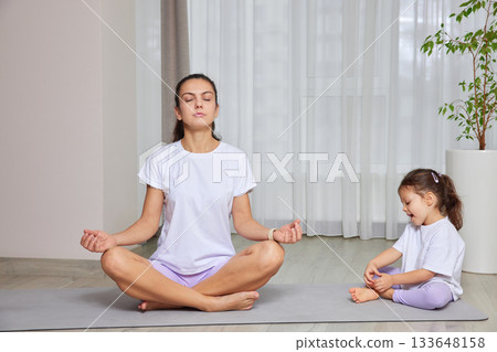 Mother and daughter sitting in a lotus pose on yoga mats at home 133648158