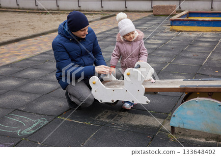 Father playing with toddler daughter on a seesaw at an outdoor playground in winter Father playing with toddler daughter on a seesaw at an outdoor playground in winter 133648402