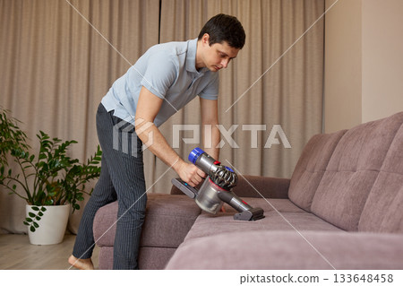 Man vacuuming sofa in living room, performing housework for maintaining home cleanliness 133648458
