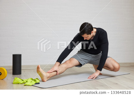Young man doing a hamstring stretch on a yoga mat at home, practicing fitness Young man doing a hamstring stretch on a yoga mat at home, practicing fitness 133648528