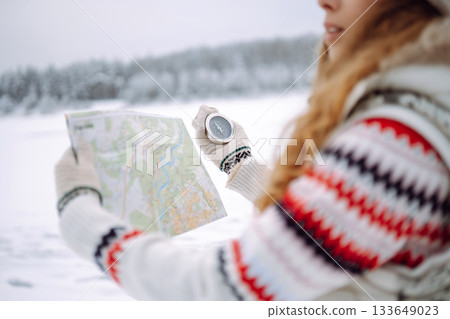 Curly-haired young woman standing in the wild with a map and compass in a snowy forest. 133649023